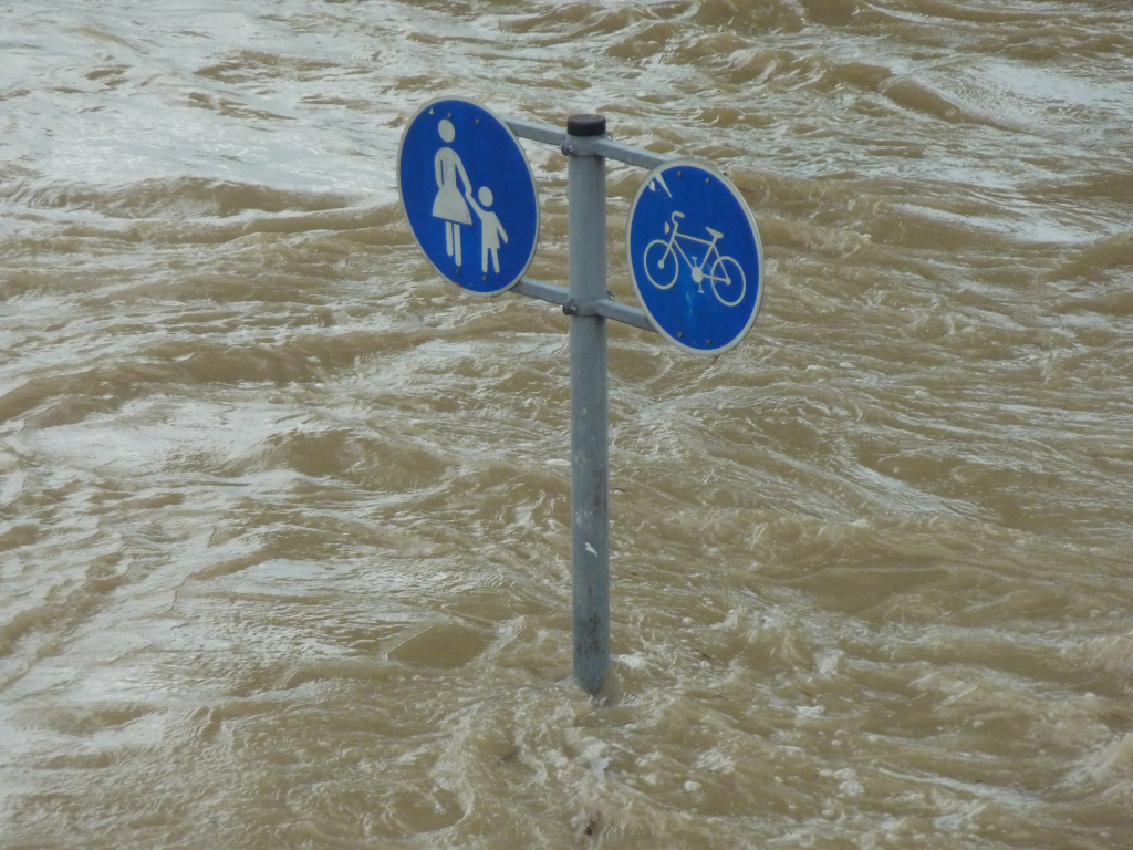Hochwasserflut, aus der ein Mast mit zwei Schildern herausragt. Es ist ein blaues Schild für Fußgänger und ein blaues für Radfahrer.